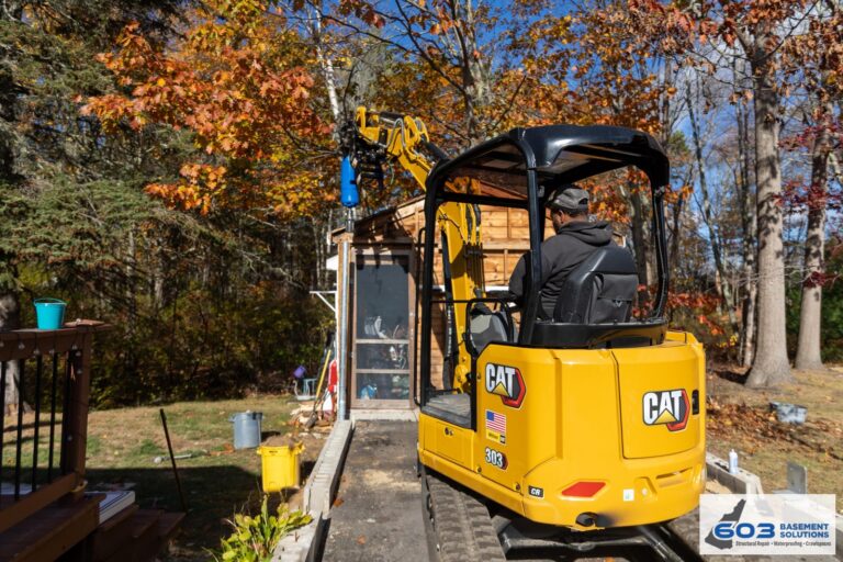 Mini-excavator with hydraulic drive head installing helical piers beside a New Hampshire home