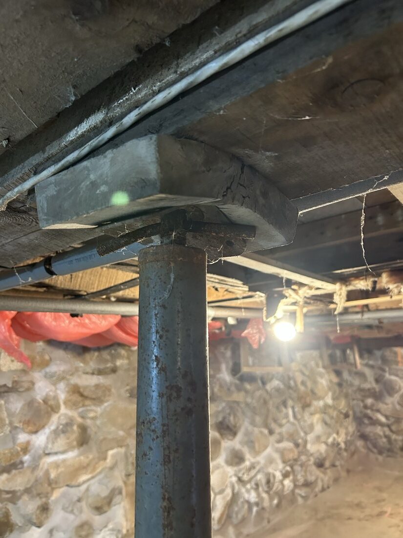 Rusting steel column under a built-up wood girder, fieldstone foundation wall behind.