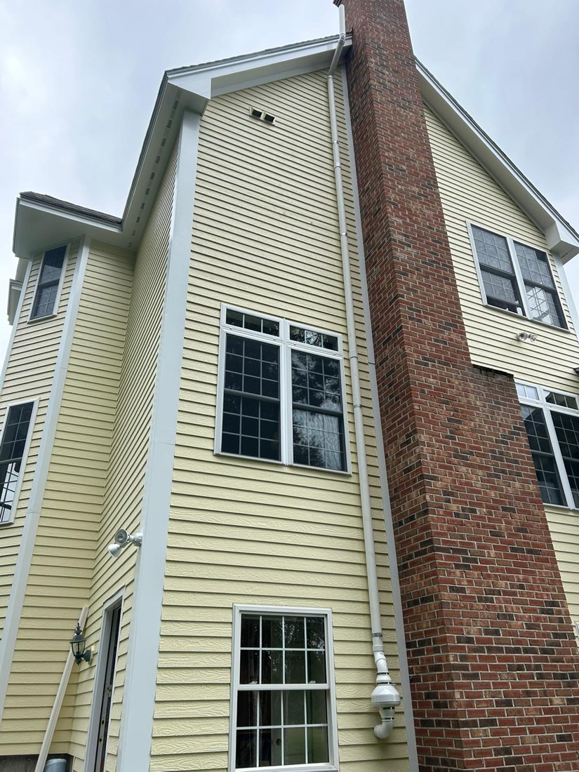 Exterior radon riser on a pale yellow clapboard house, run beside a brick chimney, discharge above the roof.